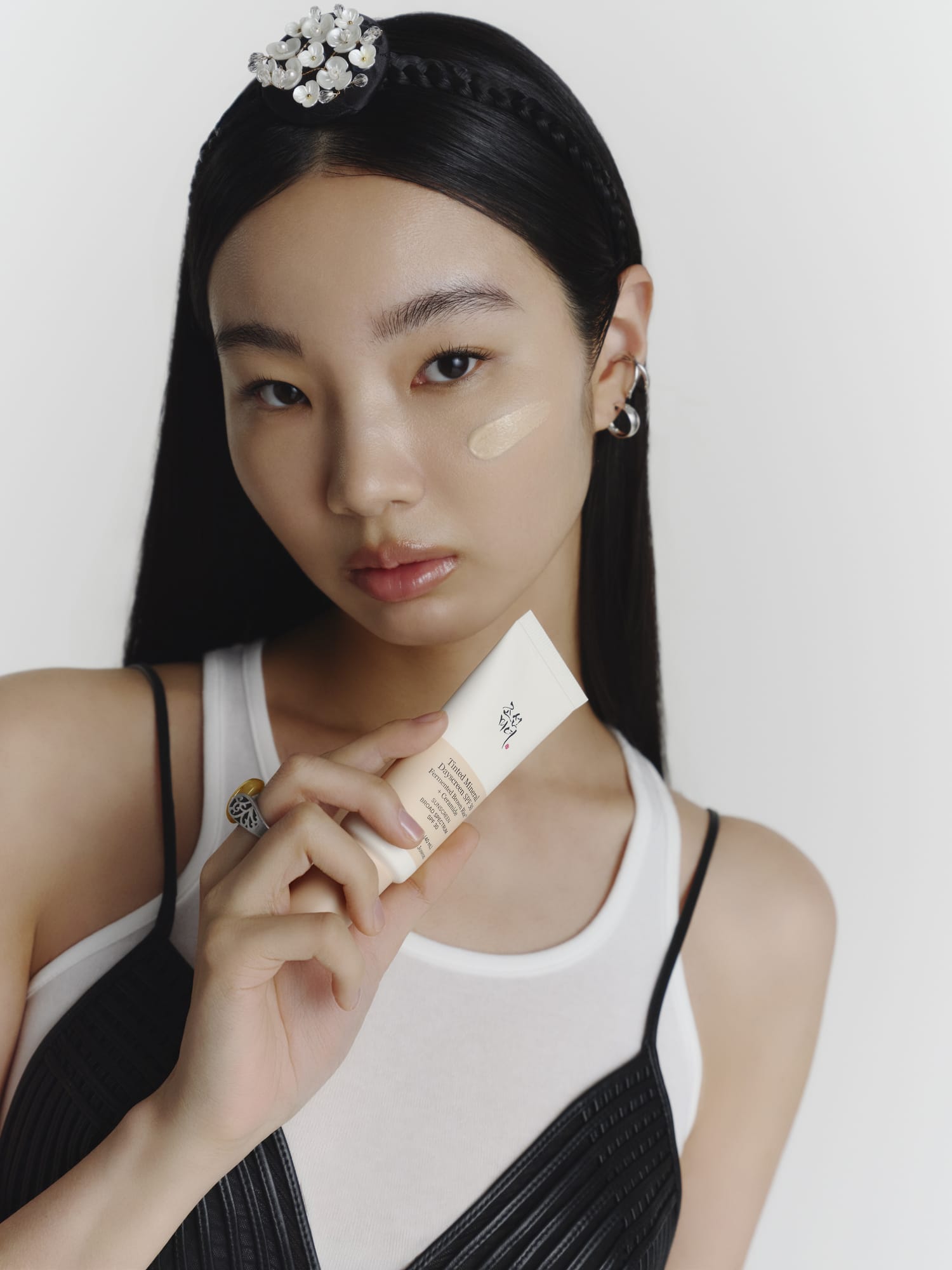 A woman with long black hair holds a Tinted Mineral Dayscreen tube near her chest while wearing a white and black top against a plain light background.