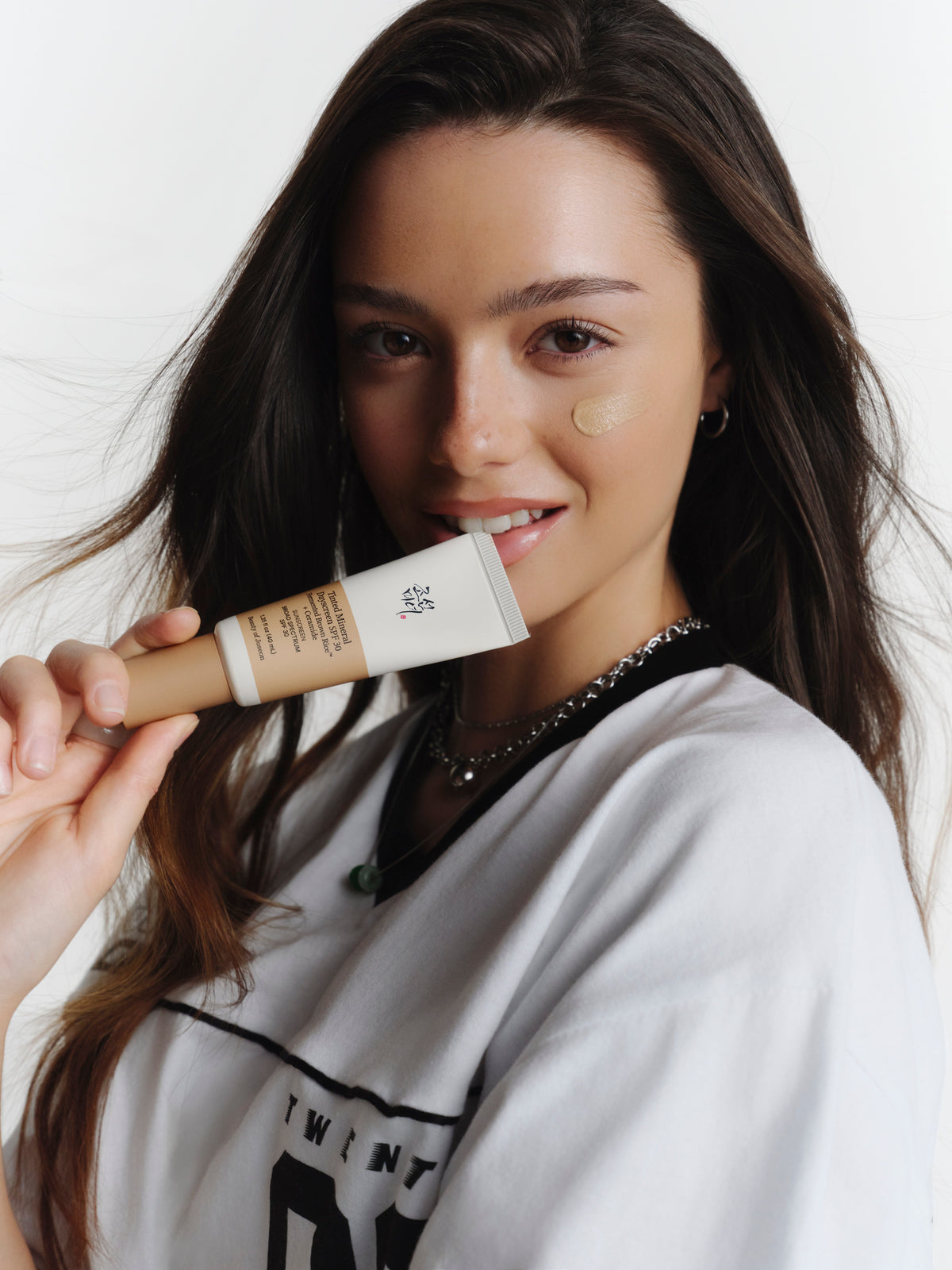 A female model smiling while holding a tube of Beauty of Joseon Tinted Mineral Dayscreen with product sample applied on her cheek.